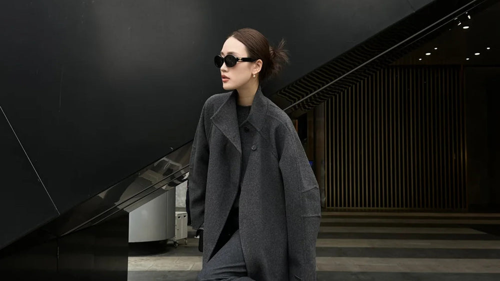 A woman in long grey outerwear beside dark contemporary architecture.