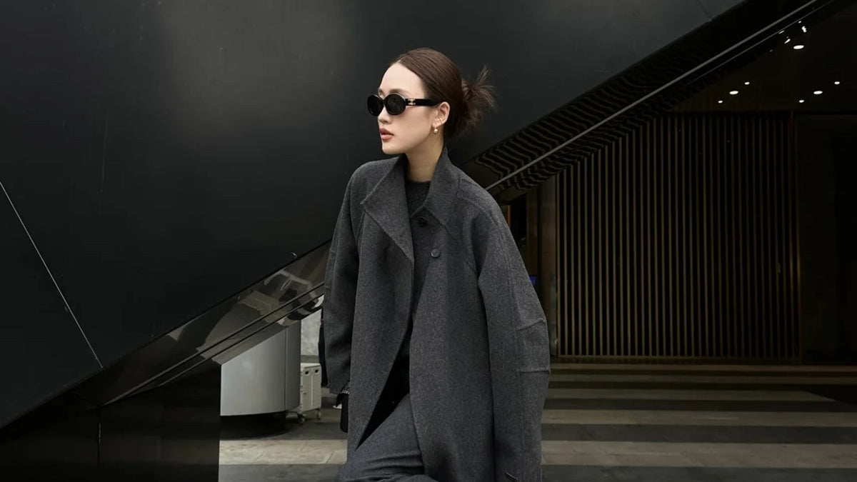 A woman in long grey outerwear beside dark contemporary architecture.
